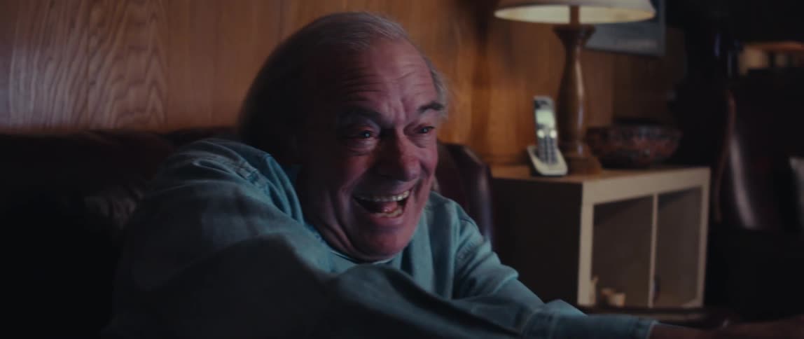 An elderly man is sitting on his couch and smiling while playing a video game