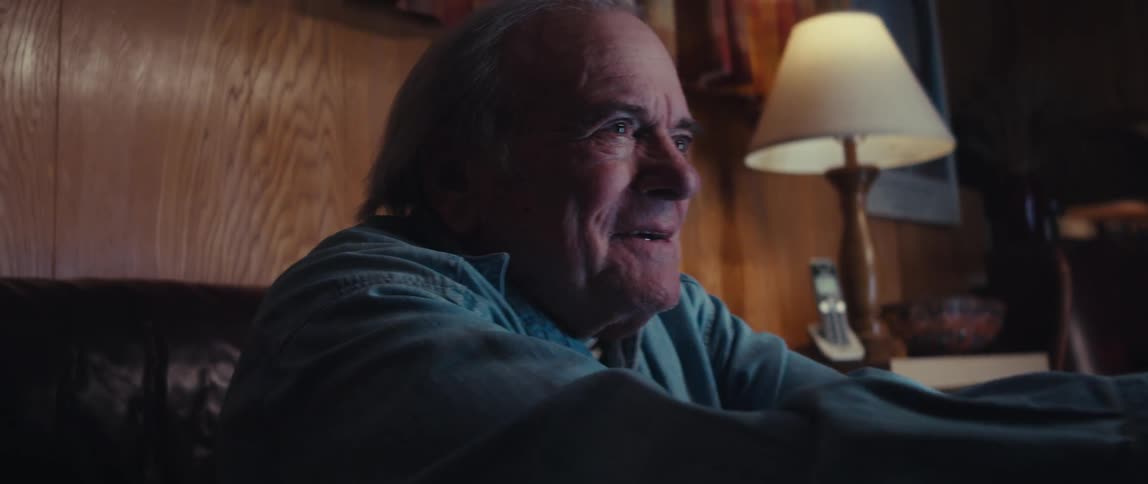 An older man sitting on a couch smiling with excitement and concentration as he plays a video game illuminated by the warm light of a nearby lamp in a cozy room