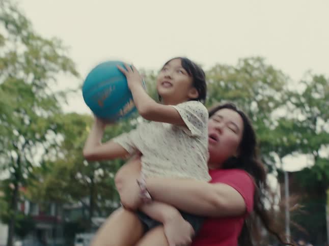 A woman picks up a little girl who is holding a blue ball and she throws the ball forward