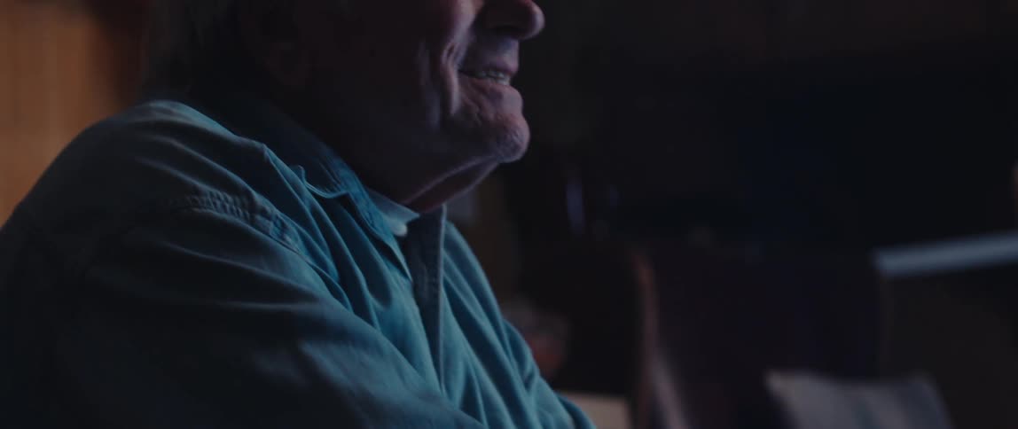 An elderly man with iron determination and a broad grin on his face plays a video game in a dimly lit room