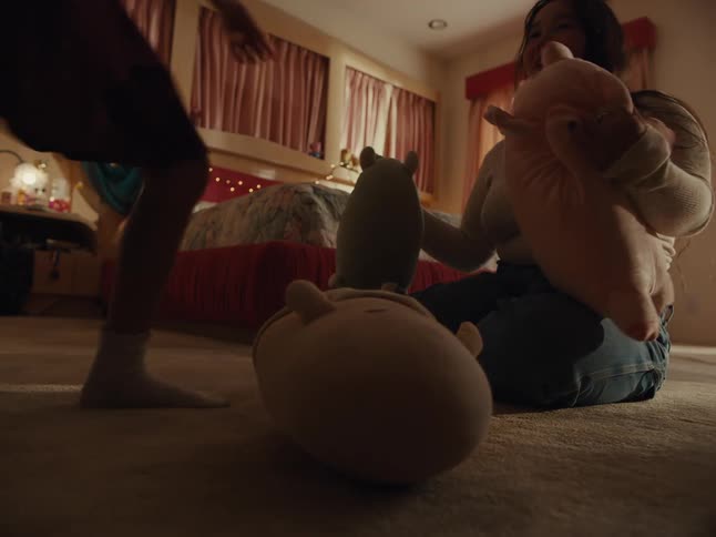 A woman sits on the floor of a bedroom as a younger person picks up plush toys and playfully interacts with her