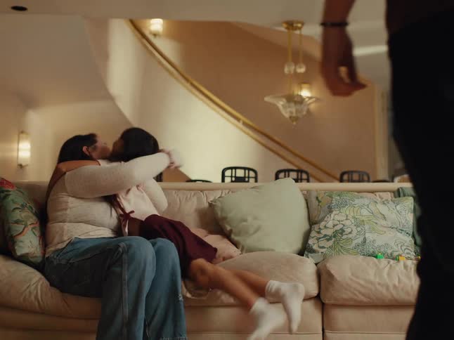 A young girl runs and hugs a woman sitting on a couch as a man walks away