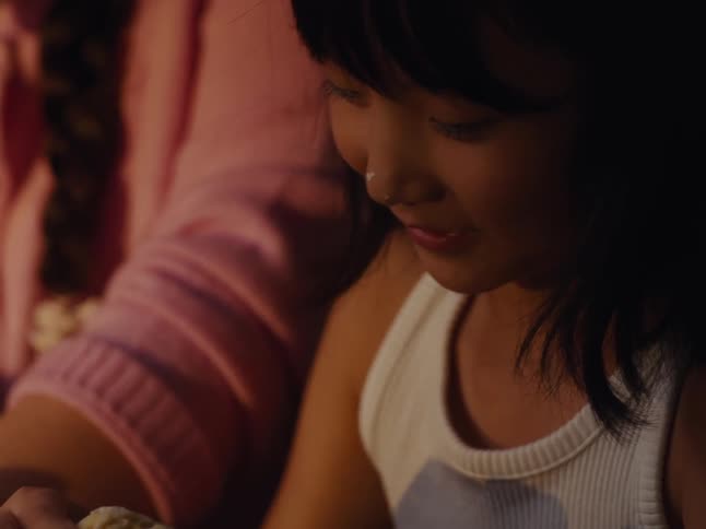 A young girl in a white tank top looks down while a slightly older person in a pink sweater laughs in the background