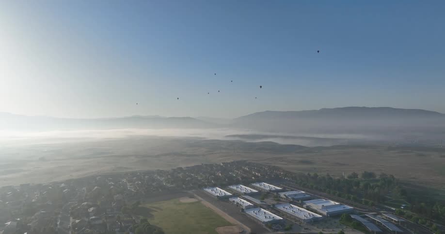 A hazy sunrise casts a soft glow over a suburban neighborhood with a sports field visible in the distance