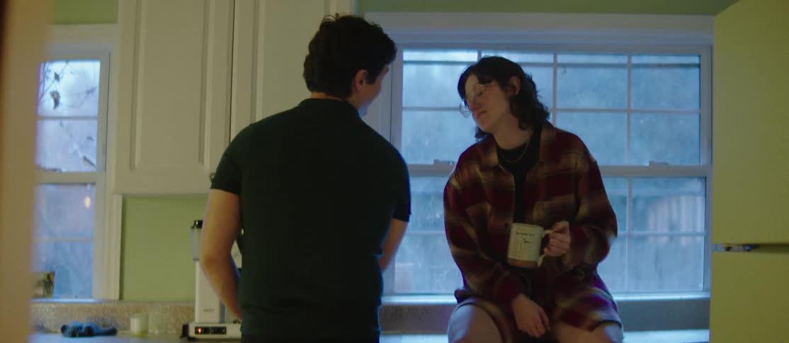 A woman holding a mug is sitting at a table talking to a man who is in front of her and she is smiling during the conversation