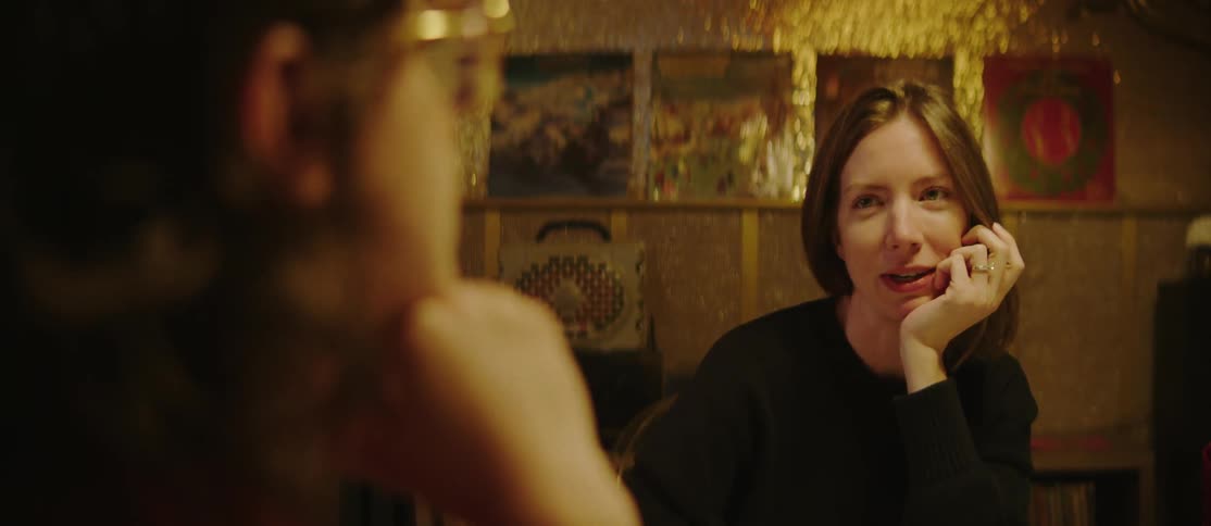 A woman with shoulder length brown hair wearing a dark sweater rests her chin on her hand and speaks to someone out of focus in a room