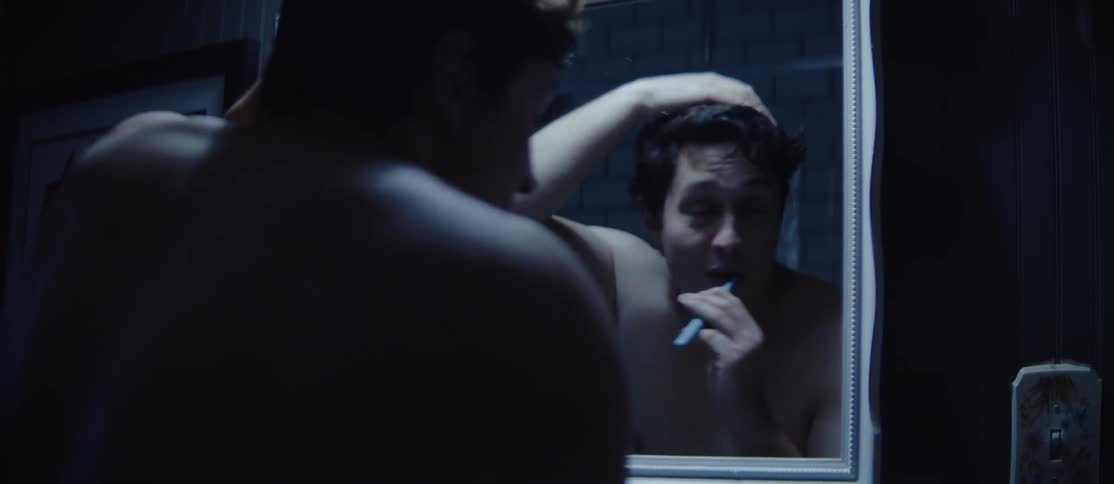 A man brushes his teeth in front of a mirror in a dimly lit bathroom