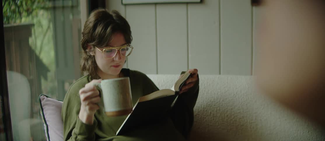 A young woman is sitting on a couch at home reading a book while drinking from a mug