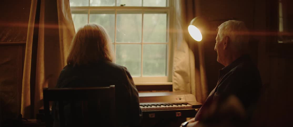Two elderly people sitting in a dimly lit room facing a window with a keyboard instrument in front of them and a lamp illuminating the scene