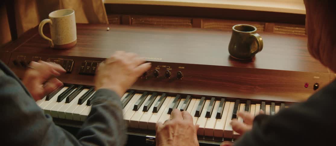 Two people sitting side by side playing an electric piano together with their hands on the keys and coffee mugs resting on the wooden surface of the instrument