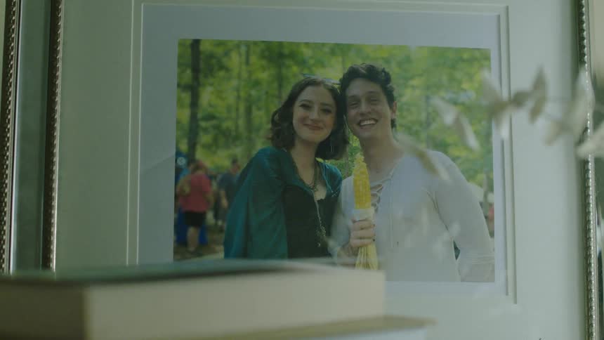 A framed photo of a smiling couple posing outdoors at a festival with one person holding a corn on the cob and trees in the background
