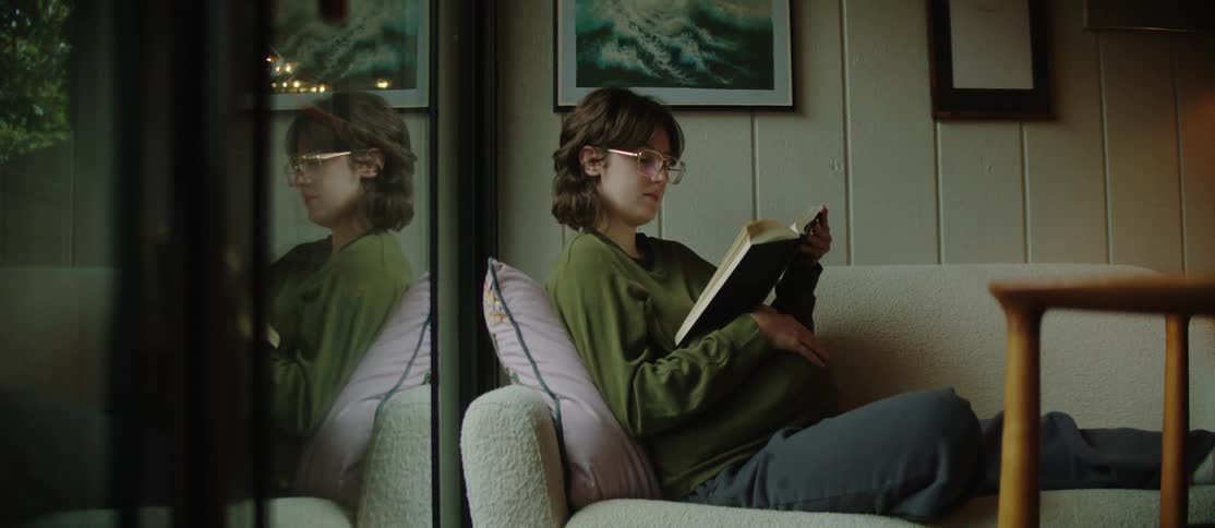 A pregnant woman with glasses and a green sweater rubs her belly while reading a book on the living room couch
