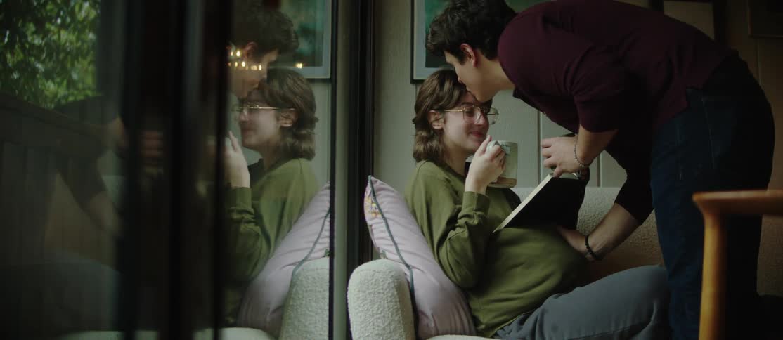 A man brings a hot beverage to his pregnant partner who is sitting on a couch at home reading a book