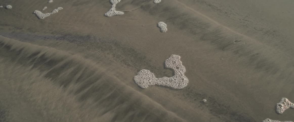Wet sand with patterns created by the receding tide and small patches of foam