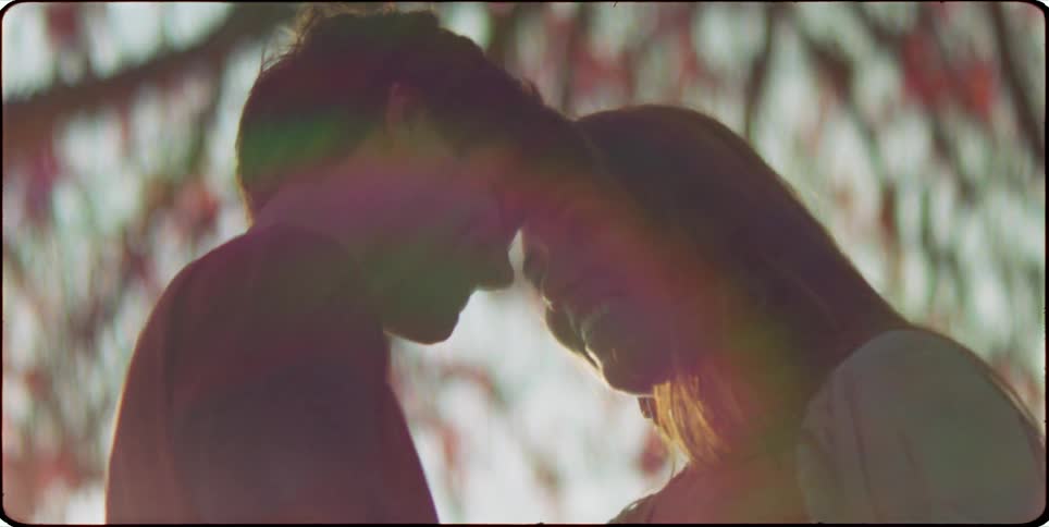 A man and woman chat and laugh drawing closer as they walk against the sunlight filtering through the tree branches