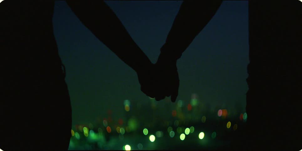A man and woman holding hands are silhouetted against a city skyline with green lights