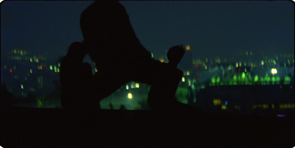 Silhouette of a person dancing while two others are sitting in the background with a cityscape at night