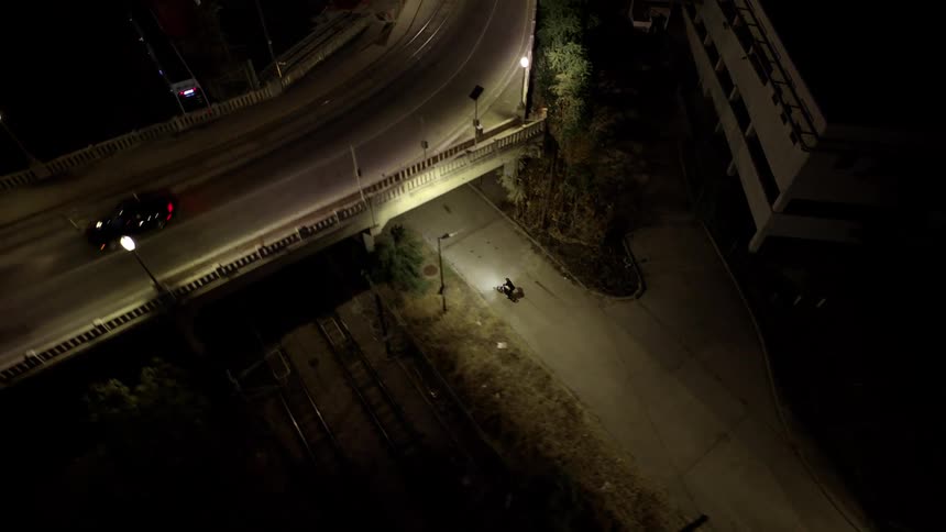 At night a cyclist rides along a paved path and the camera rises overhead for an aerial view of the path a bridge and some nearby buildings