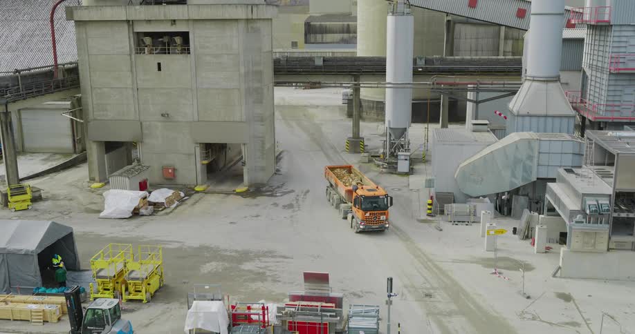 An aerial shot shows a truck carrying material driving through an industrial area with various large structures and vehicles