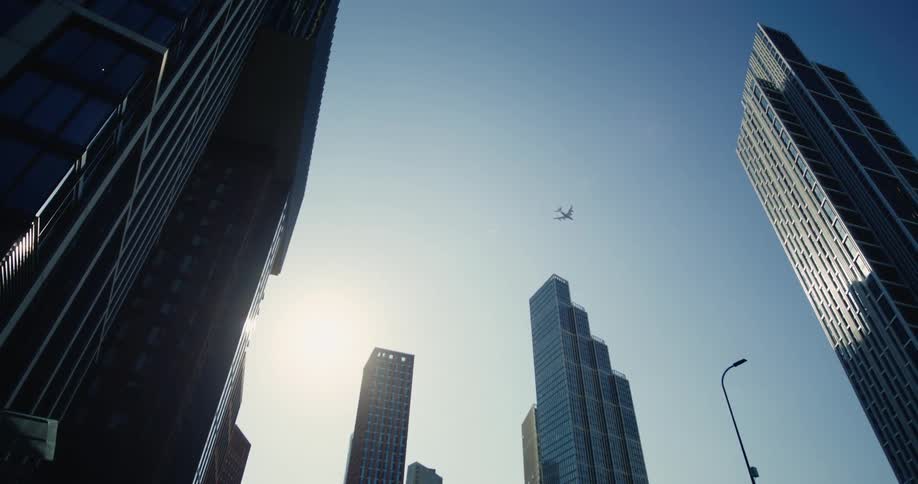 A low angle view of several modern skyscrapers against a clear blue sky with a small airplane visible in the distance