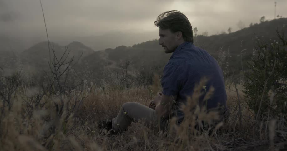 A man sitting out in a field with mountains in the background and he is staring off