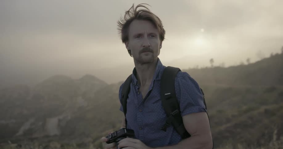A man with a backpack is standing on the edge of a mountain and he is holding a camera and turning around to see what is behind him