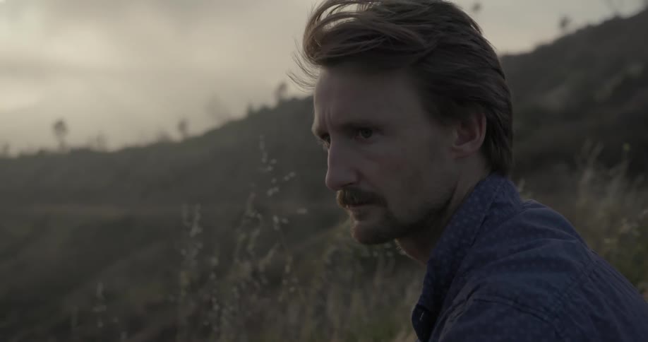 A man is sitting on a hillside and suddenly looks behind him