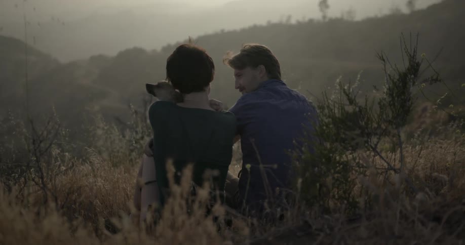 A man and woman with a small dog are sitting on a hillside and interacting