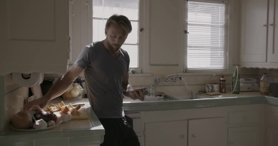 Man eating a piece of fruit while leaning on his kitchen counter and looking at his smartphone
