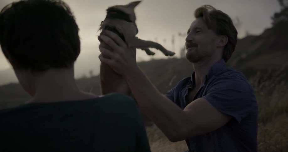 A man and a woman sitting in some tall grass and the man is holding a small dog