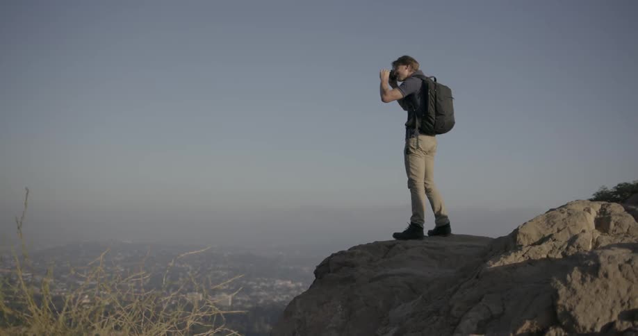 Man standing high atop  a large rock taking pictures of  what's below