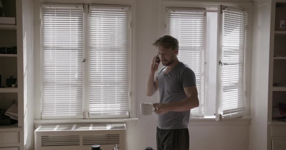 A man drinking coffee in his kitchen while talking on the phone