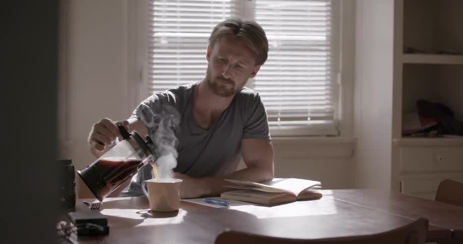 Man pours hot coffee into a mug while sitting at a table