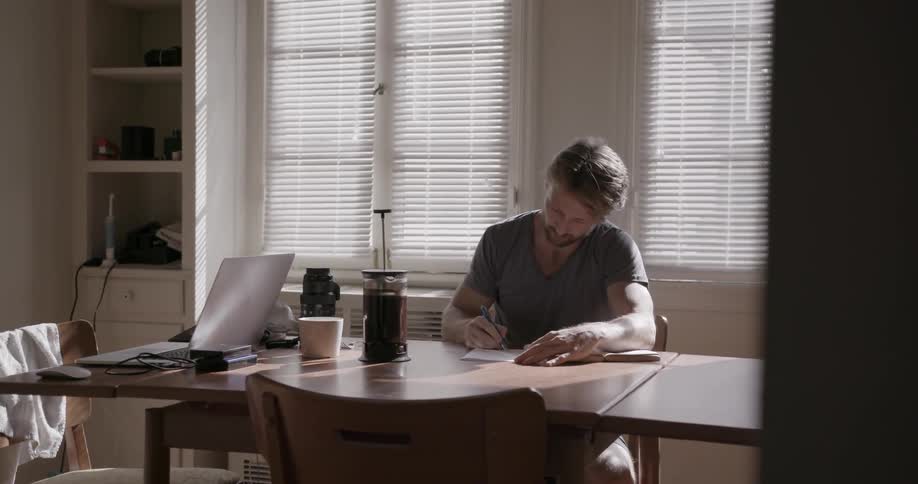 A man is sitting at a kitchen table writing in a notebook