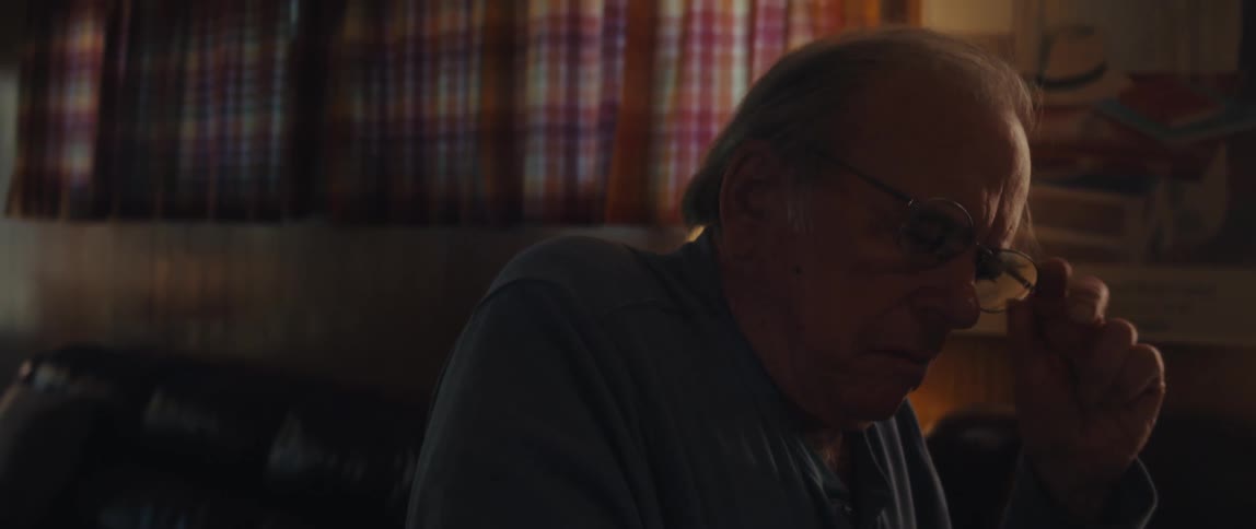 An older man with glasses wearing a dark shirt sitting in a dimly lit room takes off his glasses and looks off to the side