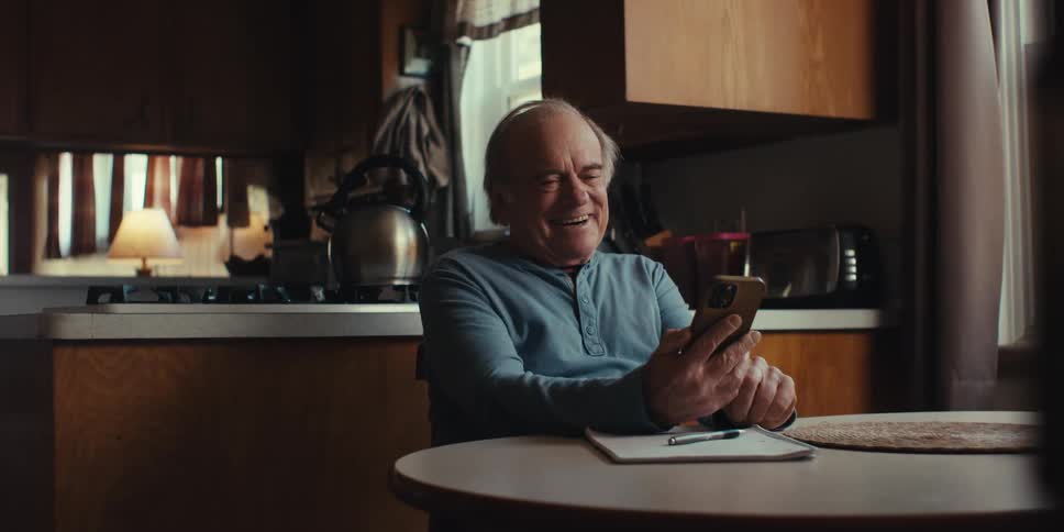 Elderly man sitting at a table laughing while interacting with a smartphone