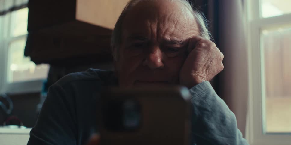 An elderly man in a blue shirt rests his head on his hand while reading his smartphone in the kitchen