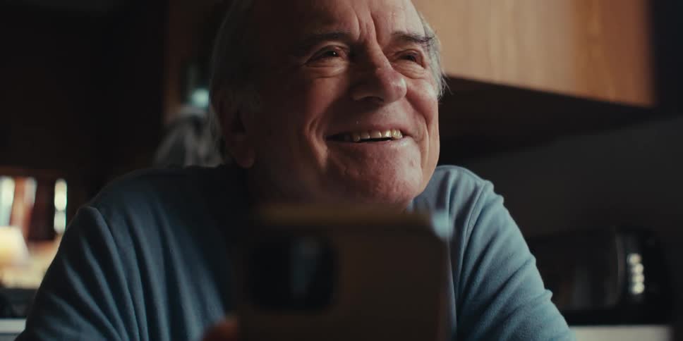 A cheerful elderly man in a blue shirt laughs while using his smartphone in the kitchen