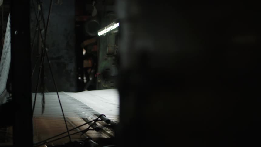 A textile loom operates in a dimly lit factory weaving white fabric