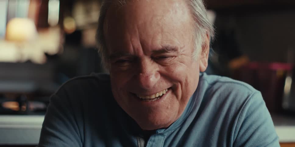 A cheerful elderly man in a blue shirt uses his smartphone with a brown case