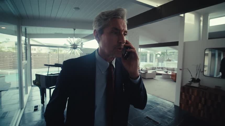 A white-haired man in a suit is standing indoors and talking on the phone