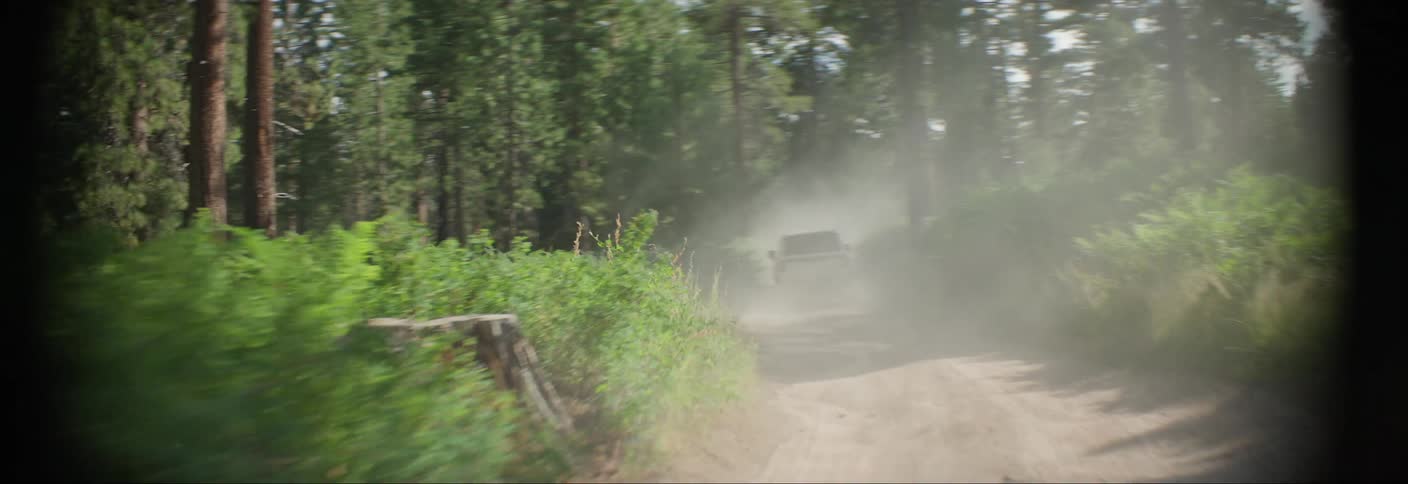 A white suv speeds down a dirt road through a dense forest kicking up a cloud of dust as it navigates the narrow path