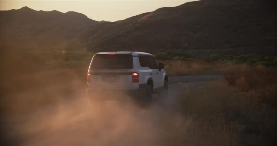 A white suv drives through a desert landscape at sunset kicking up dust and leaving a trail behind as it navigates the rugged terrain