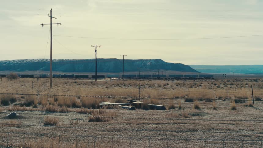 A long freight train chugs across the vast open plains dwarfed by the vast expanse of the landscape and the clear open sky