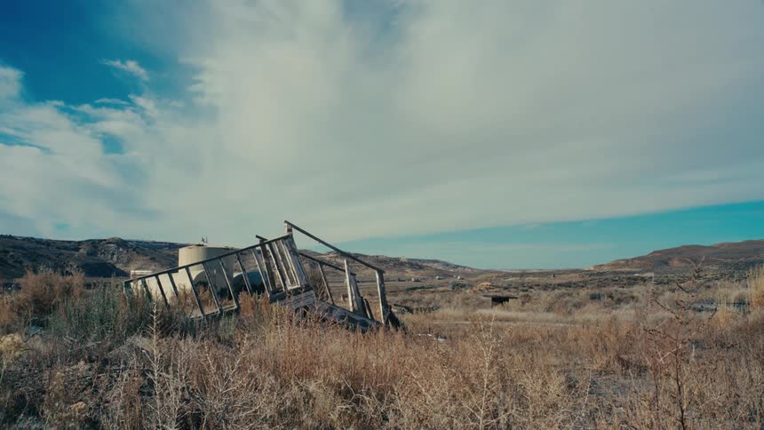 A weathered wooden structure leaning precariously in a desolate landscape hinting at a story of abandonment and the passage of time in the distance a highway with cars winds through the barren terrain