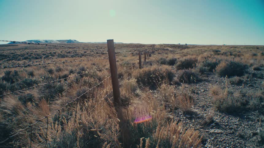 A barbed wire fence stretches across a vast open landscape with a highway and distant hills visible in the background