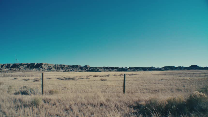 A wide shot of a vast dry grassland under a clear blue sky with rocky flattopped hills in the distance and a simple wire fence in the foreground