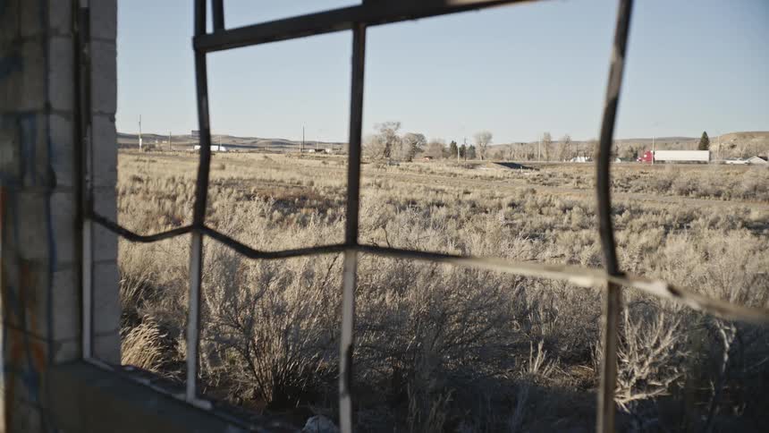 Landscape seen through a fence of sparse vegetation and a highway in the background with vehicle traffic