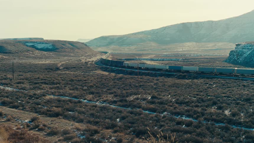 A long freight train travels through a sparsely vegetated valley with hills in the background