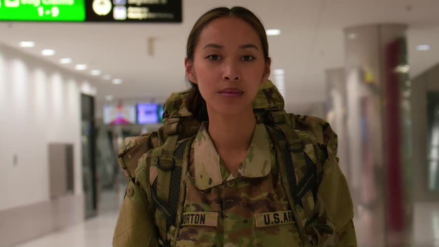 A female soldier in full military uniform carrying a backpack stands tall in a portrait taken at an airport